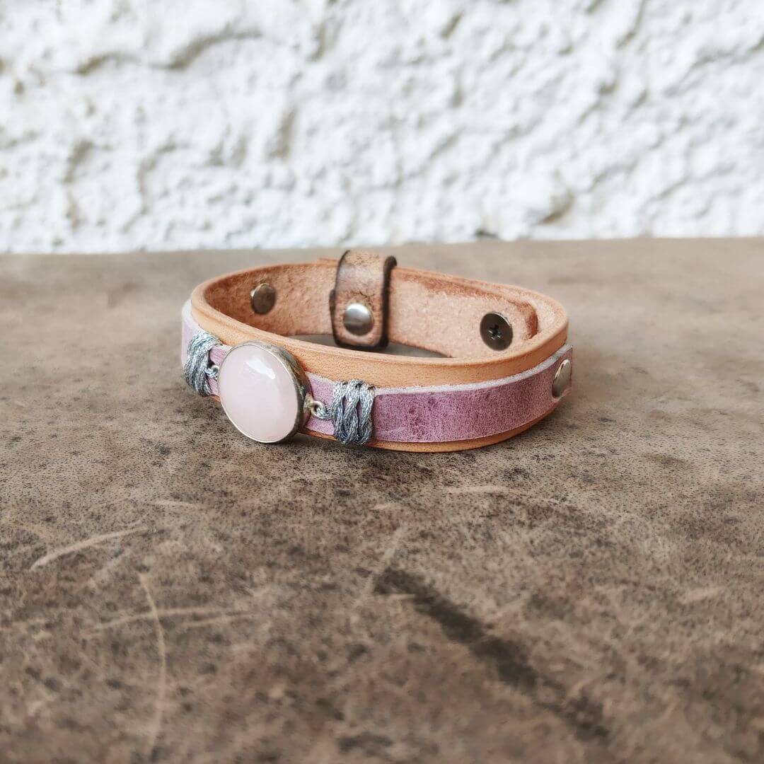 A handmade leather wrap bracelet with a pink stone cabochon and braided cord details, resting on a textured brown surface with a white stucco wall background.