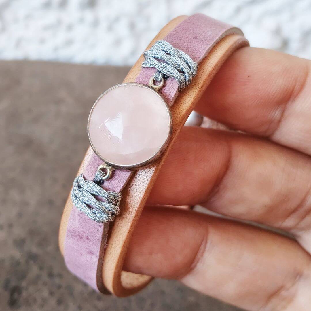A close-up of a hand holding a double-strand wrap bracelet made of dusty pink leather and light brown leather, featuring a round pale pink stone and wrapped silver cord details.