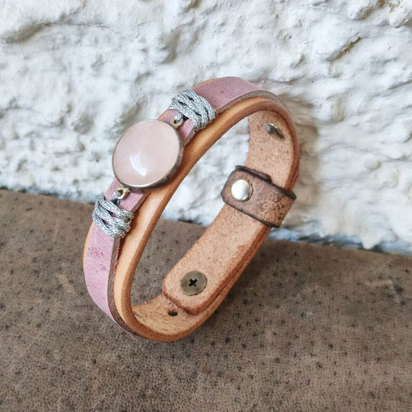 A handmade leather bracelet with a dusty pink band, a round pale pink Rose Quartz stone centerpiece, and wrapped gray cord details, resting on a textured brown surface against a white stucco wall.
