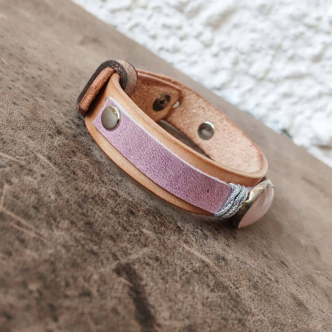 A handmade leather bracelet featuring a natural tan leather edges, silver-tone metal rivets, and a pink stone accent, resting on a textured brown surface.