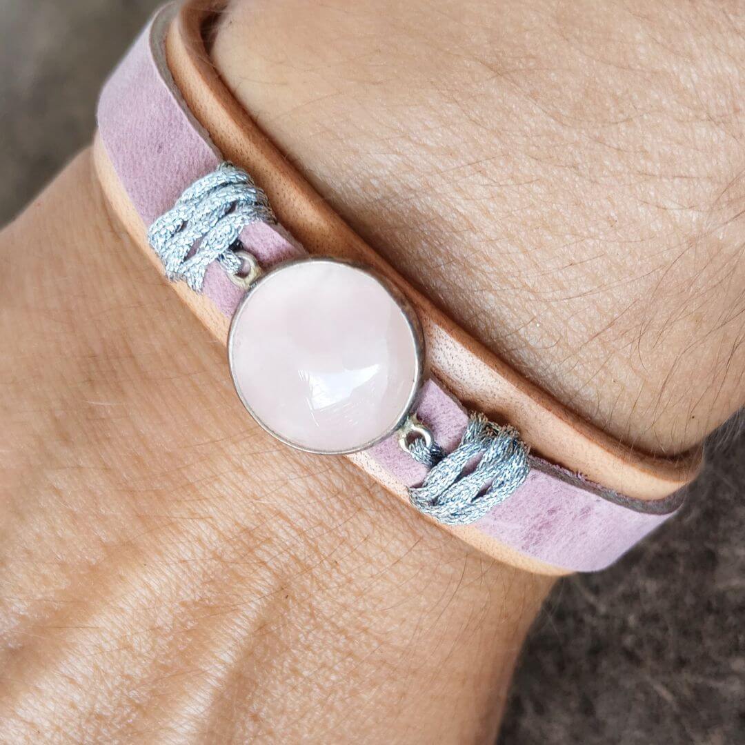 A close-up of a person's wrist wearing a double-band bracelet. The bracelet features a light pink rose quartz cabochon in a silver bezel, wrapped with a lavender suede band and a tan leather band, accented with silver braided cord.