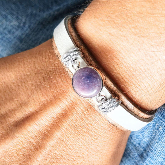 A close-up of a person's wrist wearing a layered bracelet. The bracelet features a white leather band, a tan bottom band, and a central round bezel-set purple amethyst stone.