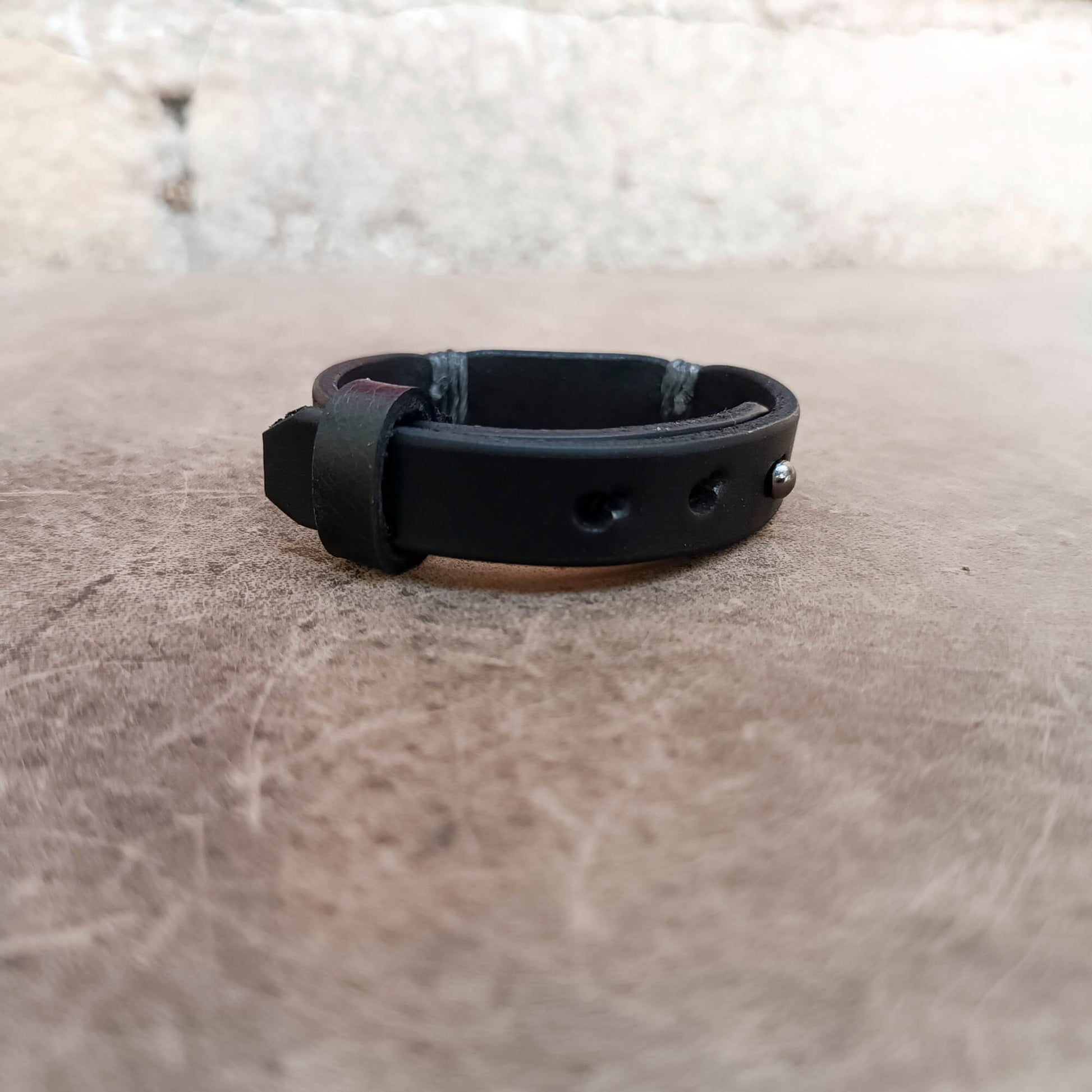 A black leather bracelet with a buckle closure and metal rivets, resting on a textured brown surface with a blurred light background.