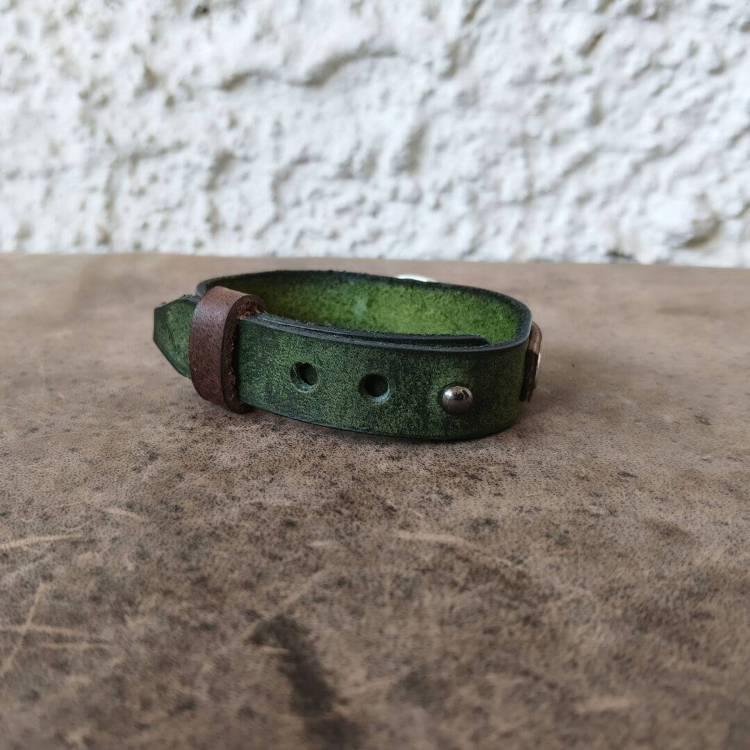 A handmade green leather cuff bracelet with a brown leather accent tab and metal rivets, resting on a textured brown surface against a white stucco wall.