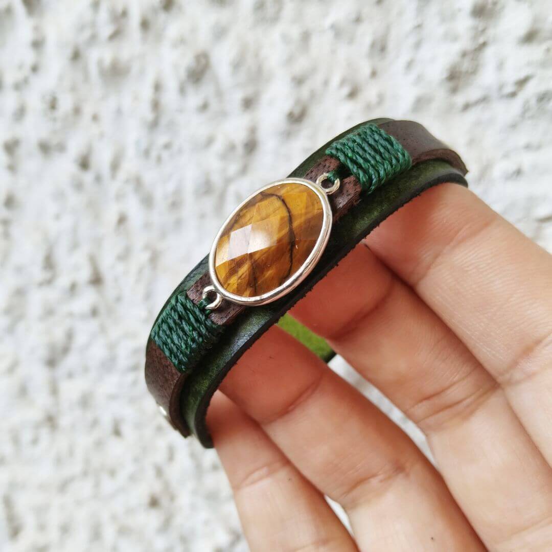 A close-up of a hand holding a wrap bracelet with a brown leather band, green braided cord, and a faceted brown tiger's eye stone in a silver bezel.