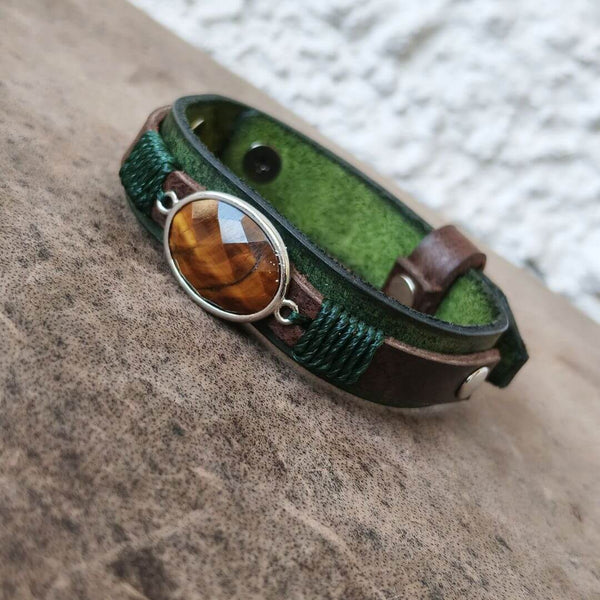 A green leather wrap bracelet with a brown leather strap, silver-toned metal hardware, and a faceted brown tiger's eye stone, displayed on a textured brown surface.