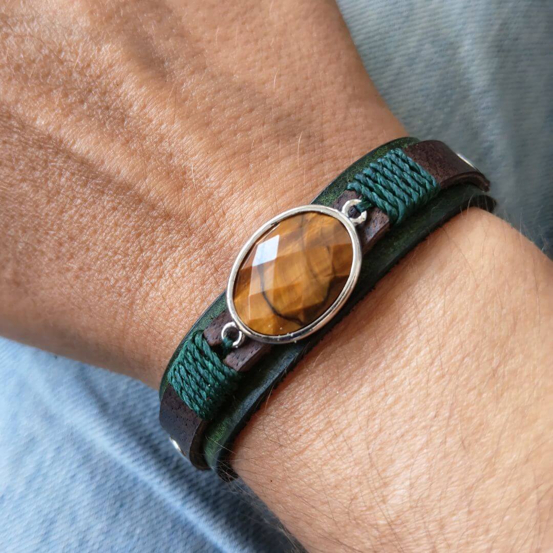 A close-up of a person's wrist wearing a multi-strand leather wrap bracelet. The bracelet features dark brown leather, a dark green braided cord, and a central oval bezel-set brown tiger's eye stone with a silver-toned metal frame.