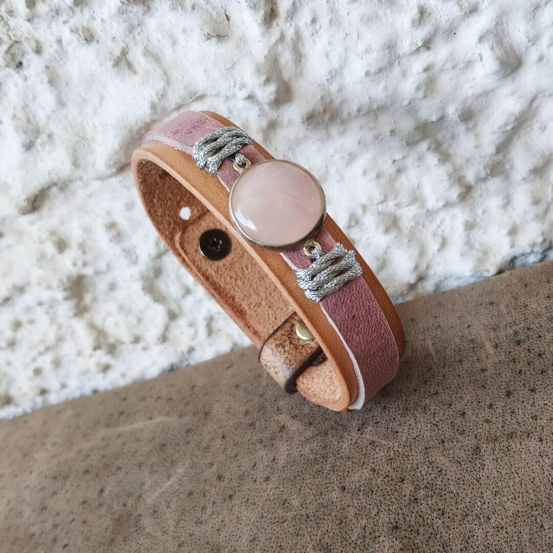 A handmade leather cuff bracelet with a dusty pink stone cabochon and silver-toned metal accents, resting on a textured brown surface against a white stucco wall.
