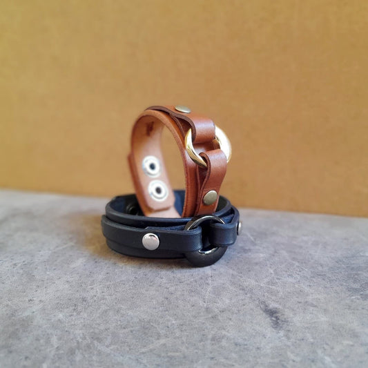 The Personalized Cremation Urn Leather Cuff: One black and one brown full-grain leather bracelet featuring a prominent stainless steel urn and metal rivets.