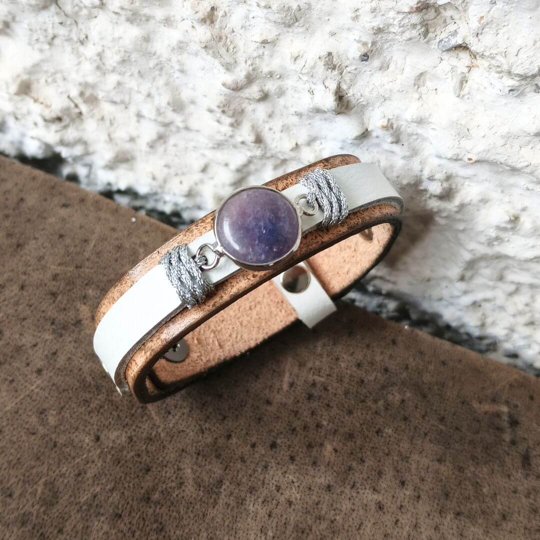 A handmade wrap bracelet featuring natural brown leather, a white leather stripe, silver gray cord wraps, and a central purple stone, displayed on a textured brown surface against a white stone background.