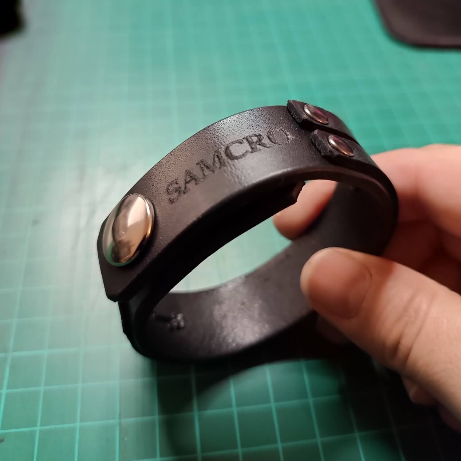 Black leather bracelet with 'SAMCRO' engraving held by a hand on a green cutting mat.