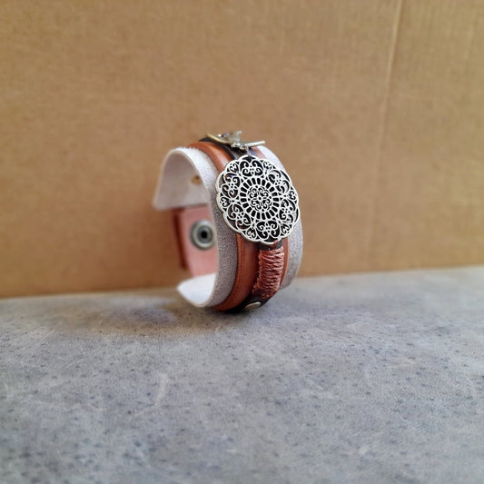 side full view of the antique bronze mandala flower motif and the little bird charm on the "Happy Beige" Mandala Cuff, showing the hand-stitched brown details.