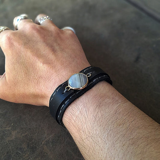 The Obsidian Cuff: An OOAK men's black leather bracelet worn on wrist, featuring a gold-plated agate cabochon with light and dark gray veins, and deep gray hand-stitched details.
