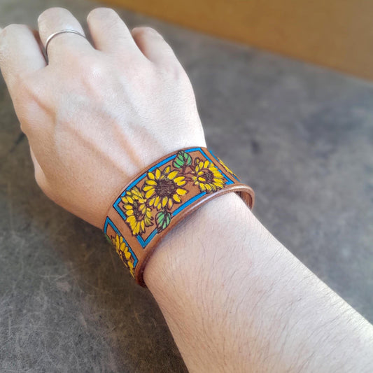 The Sunshine Bloom Leather Cuff Bracelet worn on a wrist, showcasing its vibrant hand-painted sunflower design and mustard brown Crazy Horse leather, adding a Western, joyful touch.