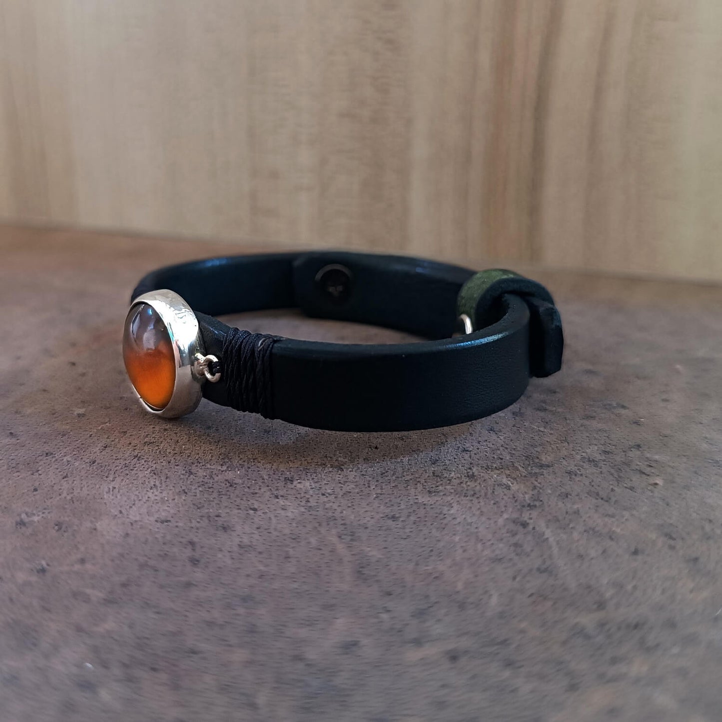 Black leather bracelet with a sterling silver Tiger's Eye pendant on a textured surface, side view