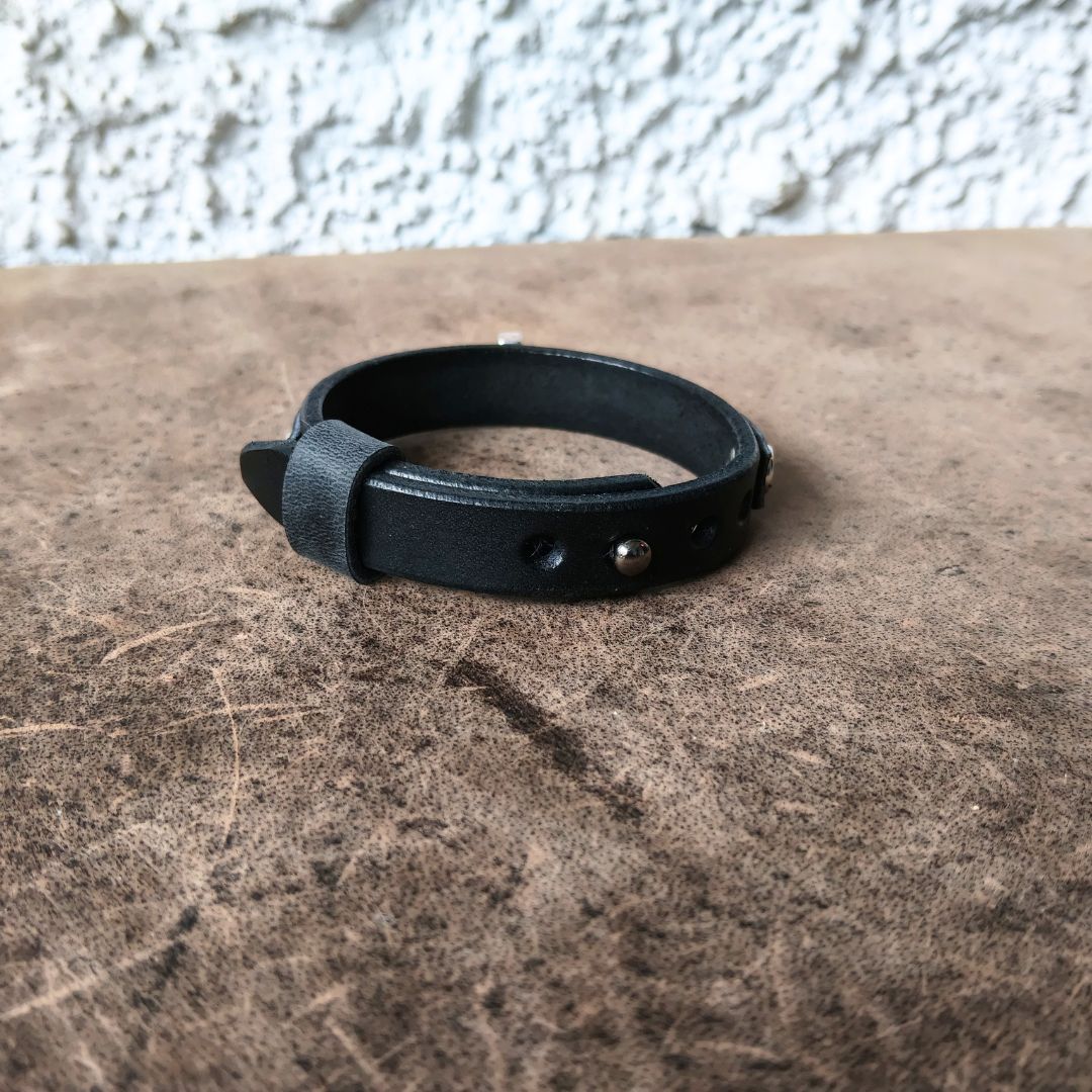 A black leather adjustable cuff bracelet with silver-tone metal rivets and a buckle, resting on a textured brown surface against a white stucco wall.