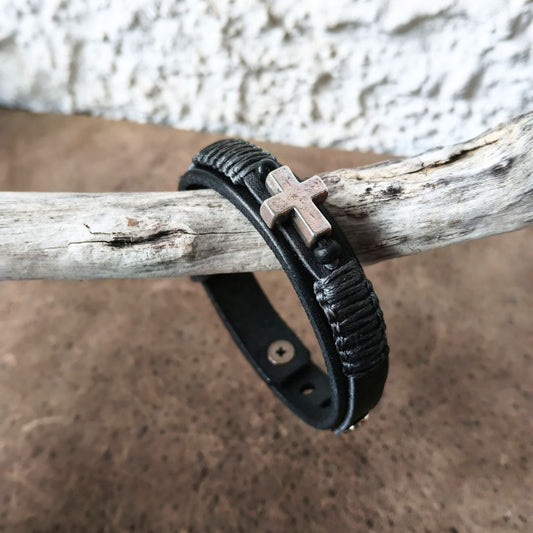 A black leather bracelet with a textured wrap design and a small metallic cross charm, resting on a weathered branch against a rustic background.