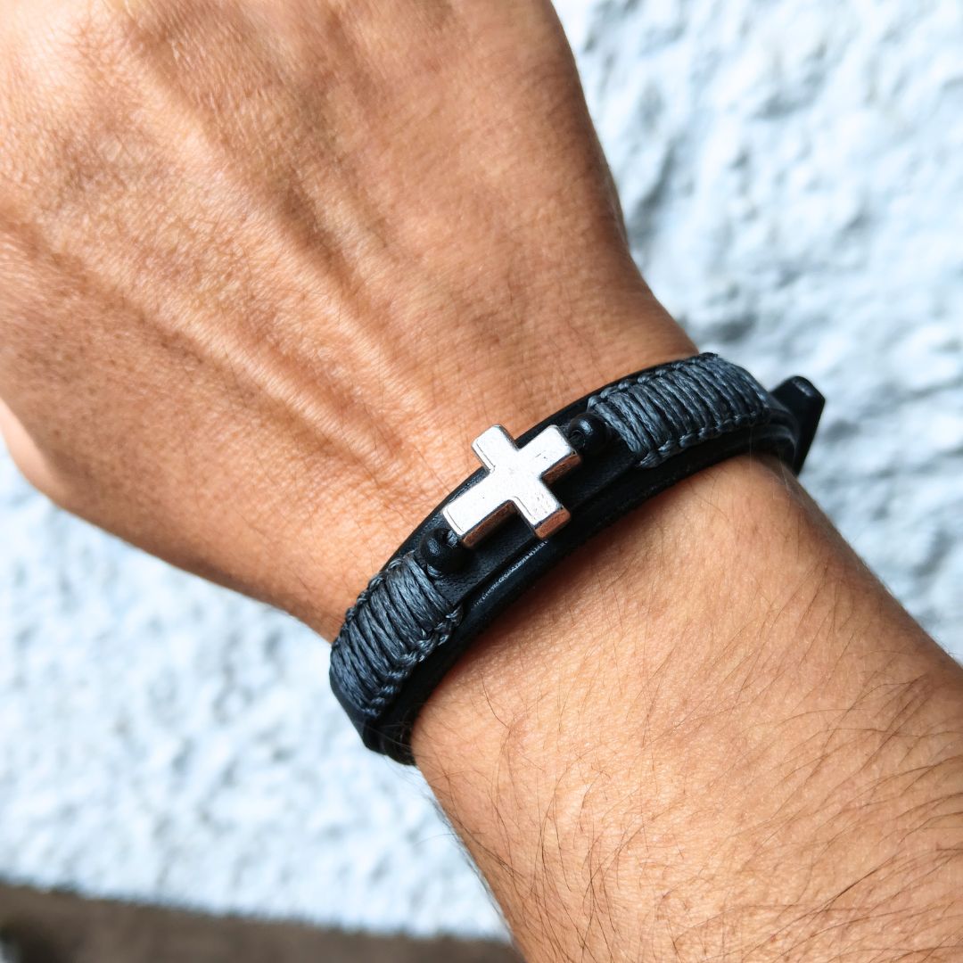A close-up of a person's wrist wearing a black braided leather bracelet with a silver-toned cross charm.