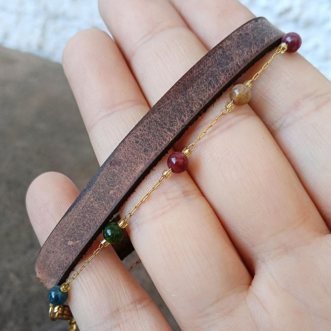 A close-up of a hand holding a brown leather bracelet with a thin gold chain and small multicolored beads.