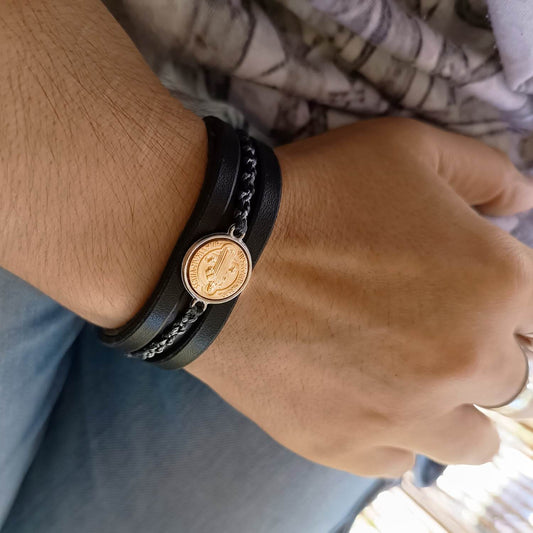 Close-up of a wrist wearing a black leather bracelet with a gold and silver Byzantine style coin pendant.