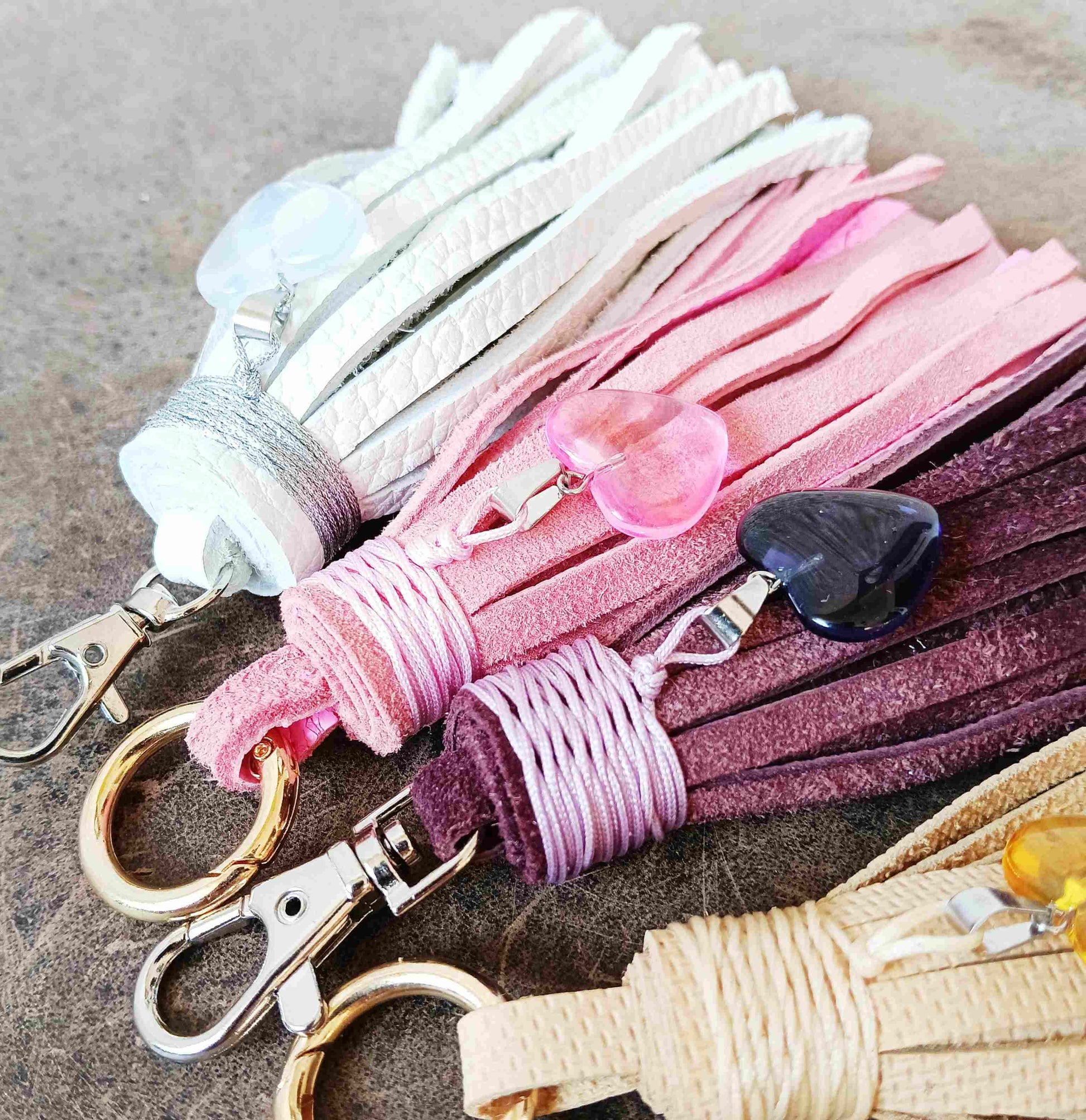 Four leather tassel keychains in white, pink, mauve, and beige, arranged diagonally on a textured gray surface. Each keychain features a metal lobster clasp, a braided cord wrap, and a decorative stone charm.
