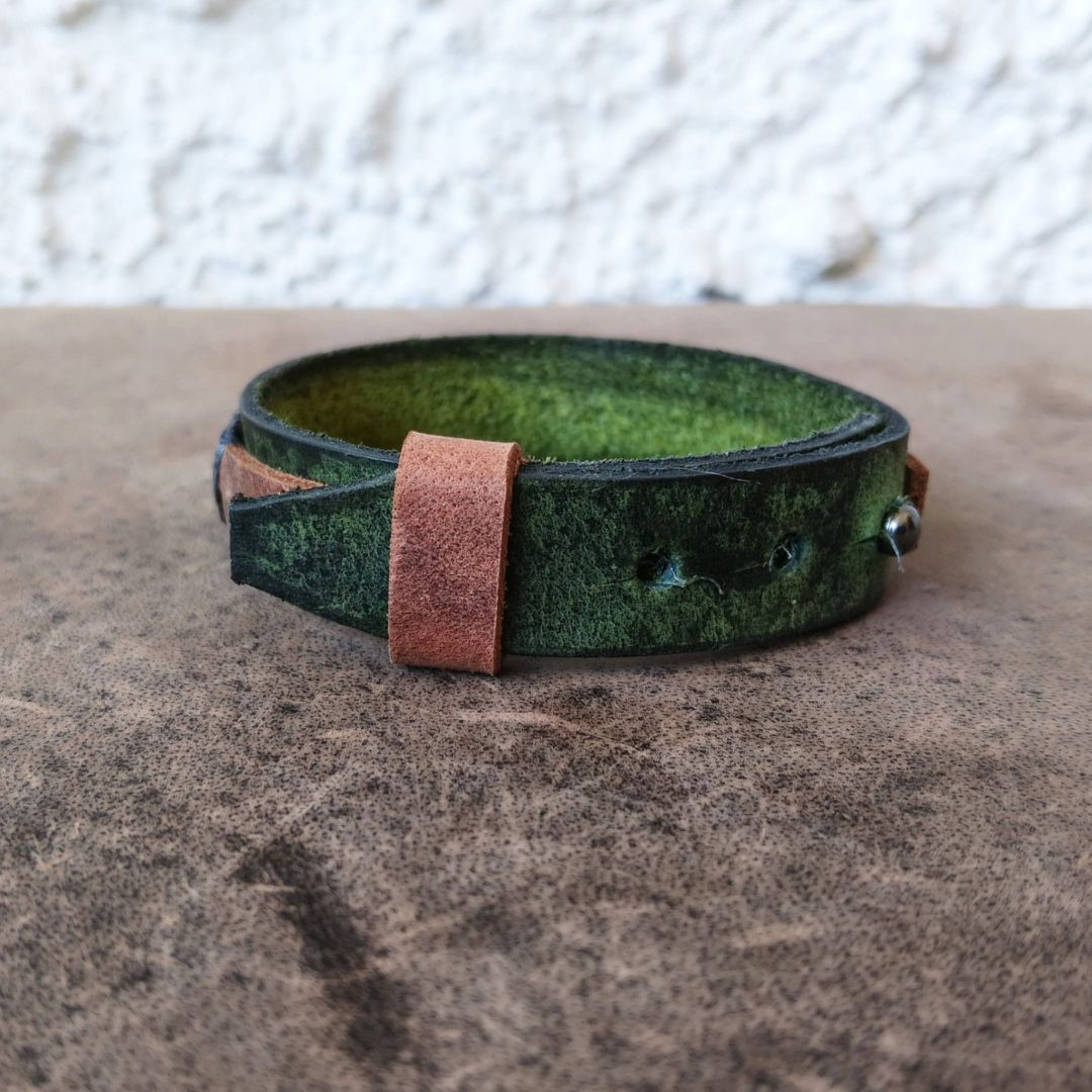 A handmade green leather wrap bracelet with a contrasting brown leather tab and silver metal snap closure, resting on a textured brown surface.