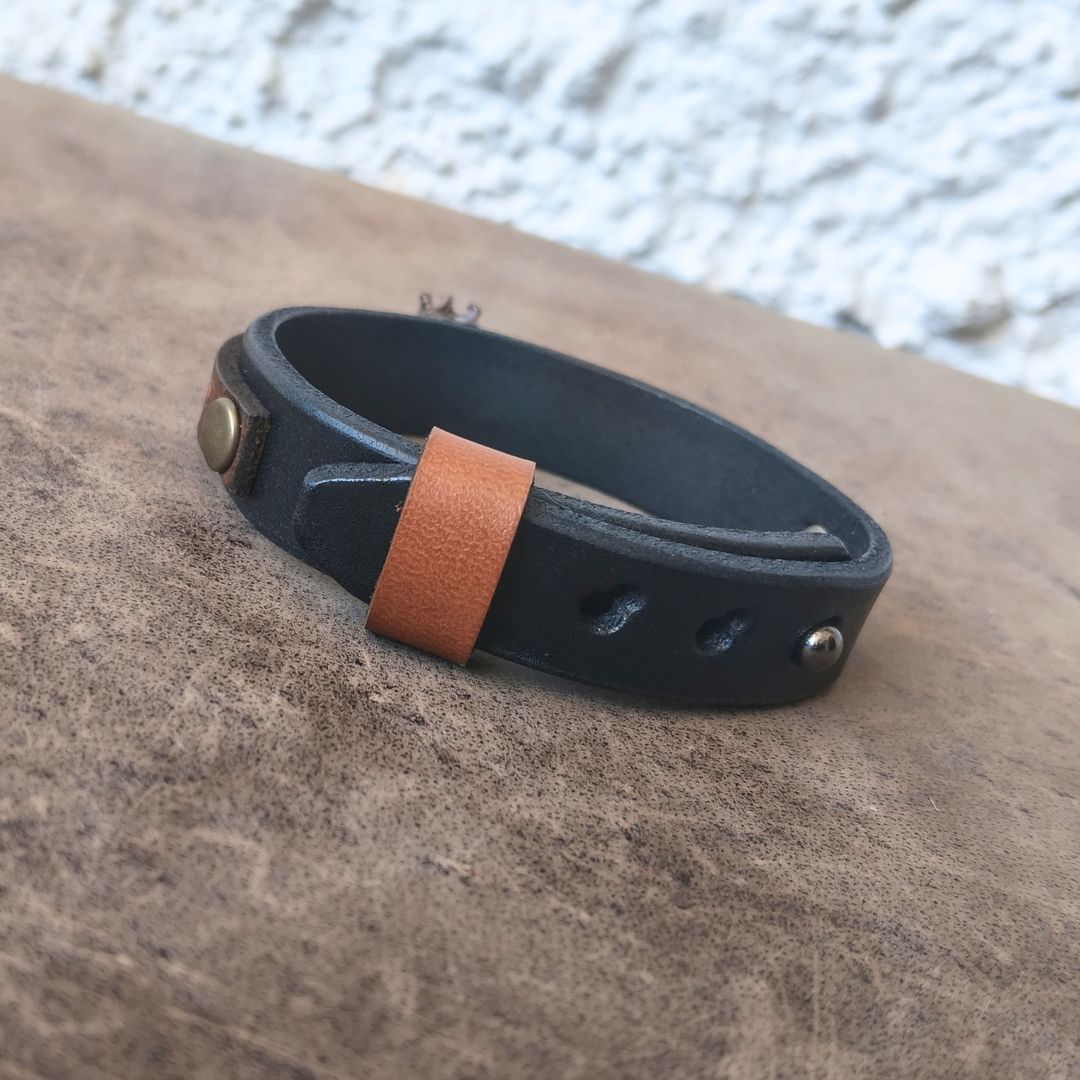 A black leather bracelet with a contrasting tan leather tab and brass-tone rivets, resting on a textured brown surface.
