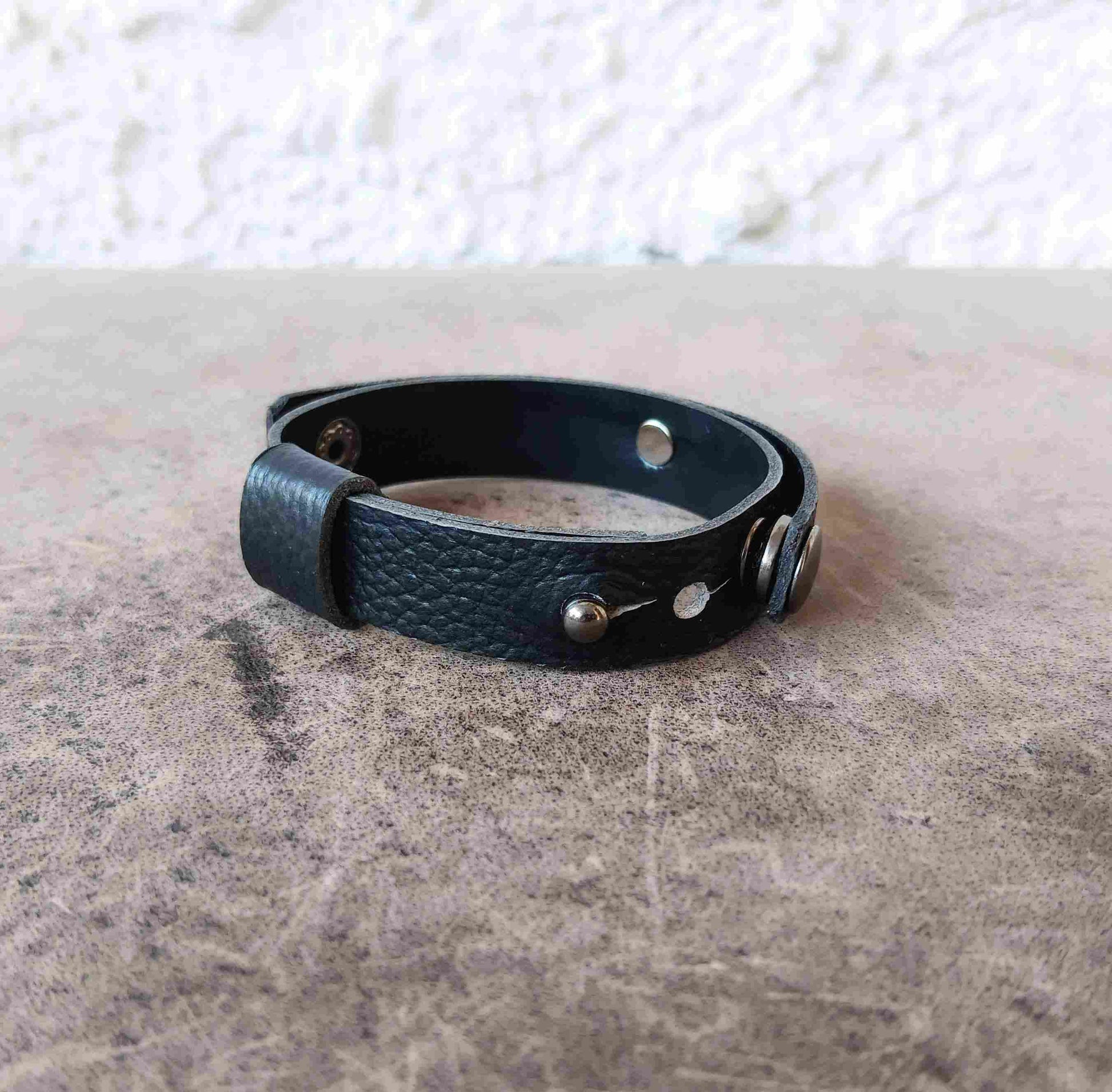 A black textured leather cuff bracelet with silver-tone metal rivets and a toggle clasp, resting on a textured gray surface.