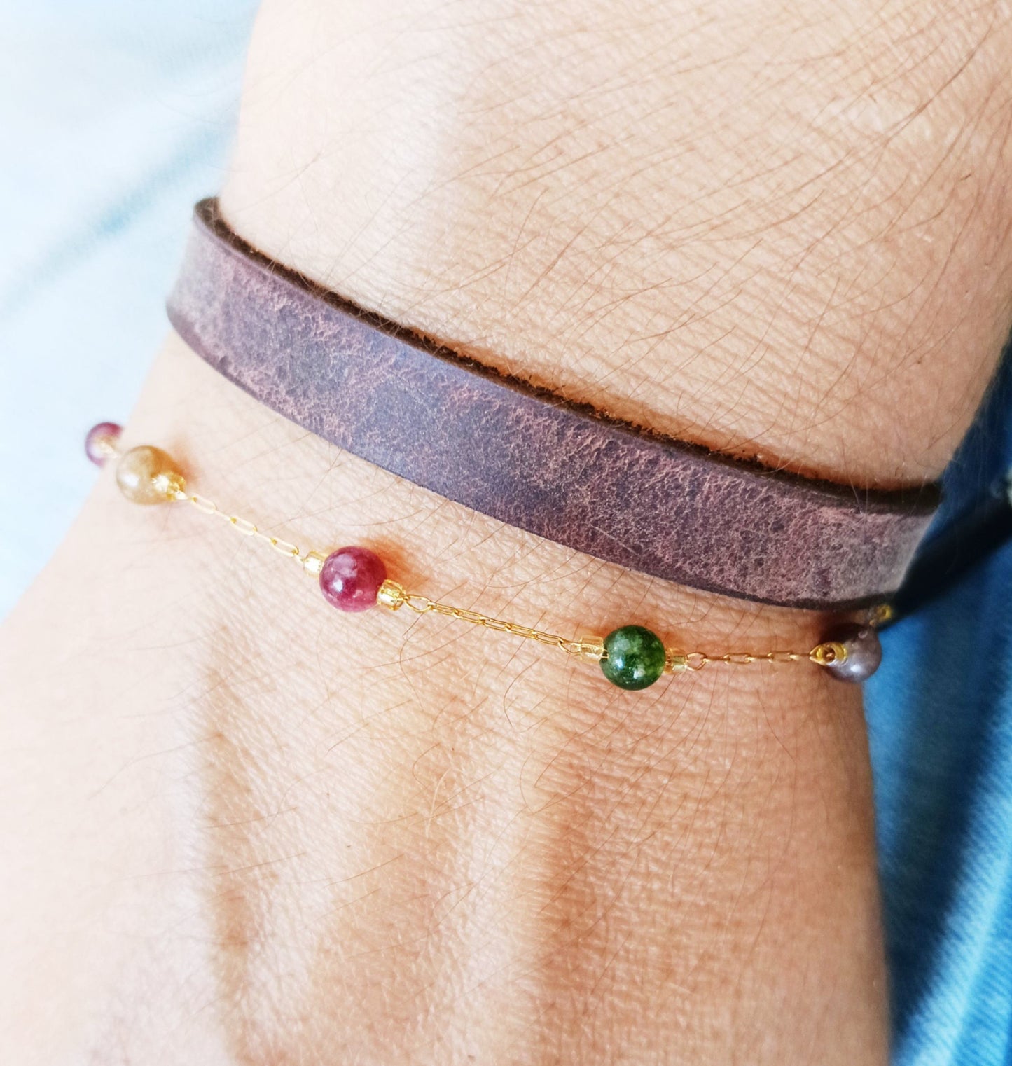 A close-up of a person's wrist wearing two bracelets: a wide dark brown leather wrap bracelet and a delicate gold chain bracelet with small multicolored beads.