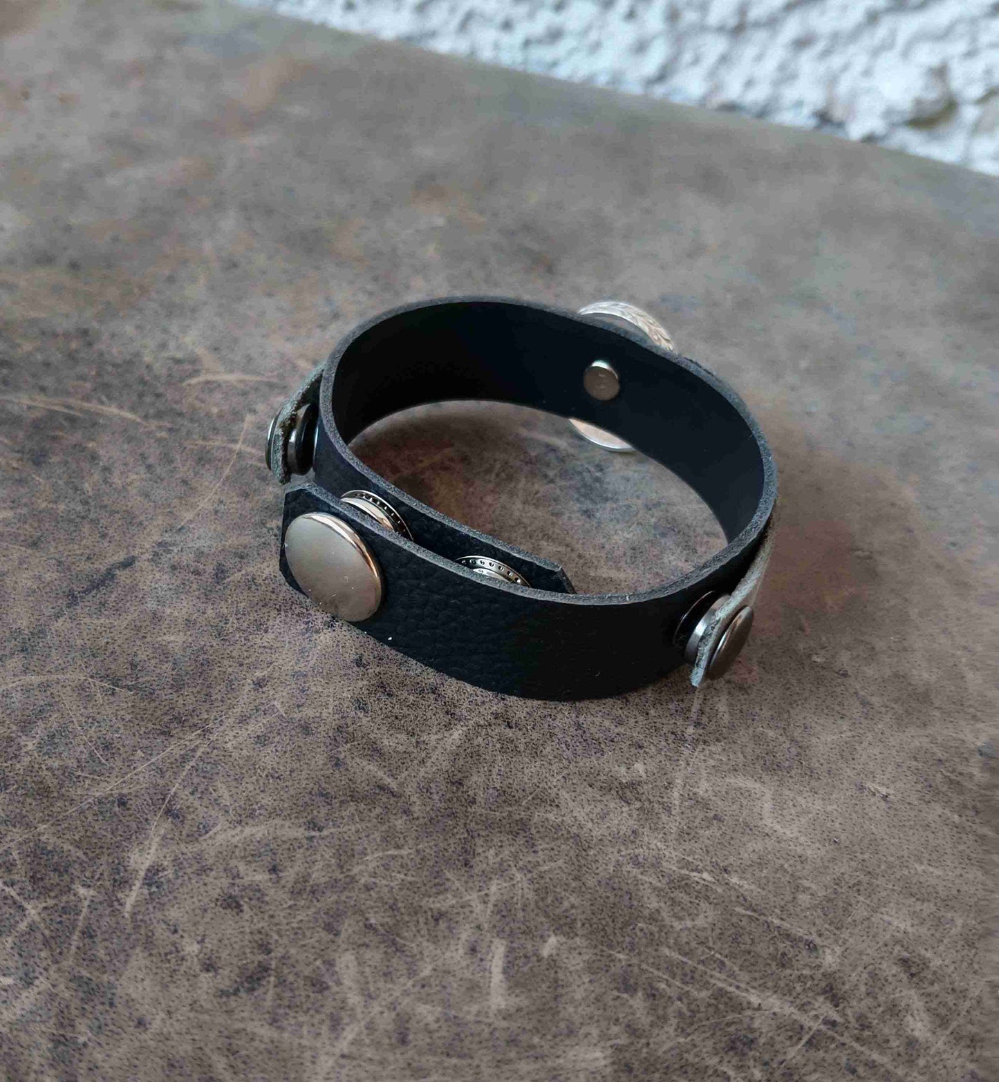 A black textured leather cuff bracelet with silver-toned metal rivets and snap buttons, resting on a textured brown surface.