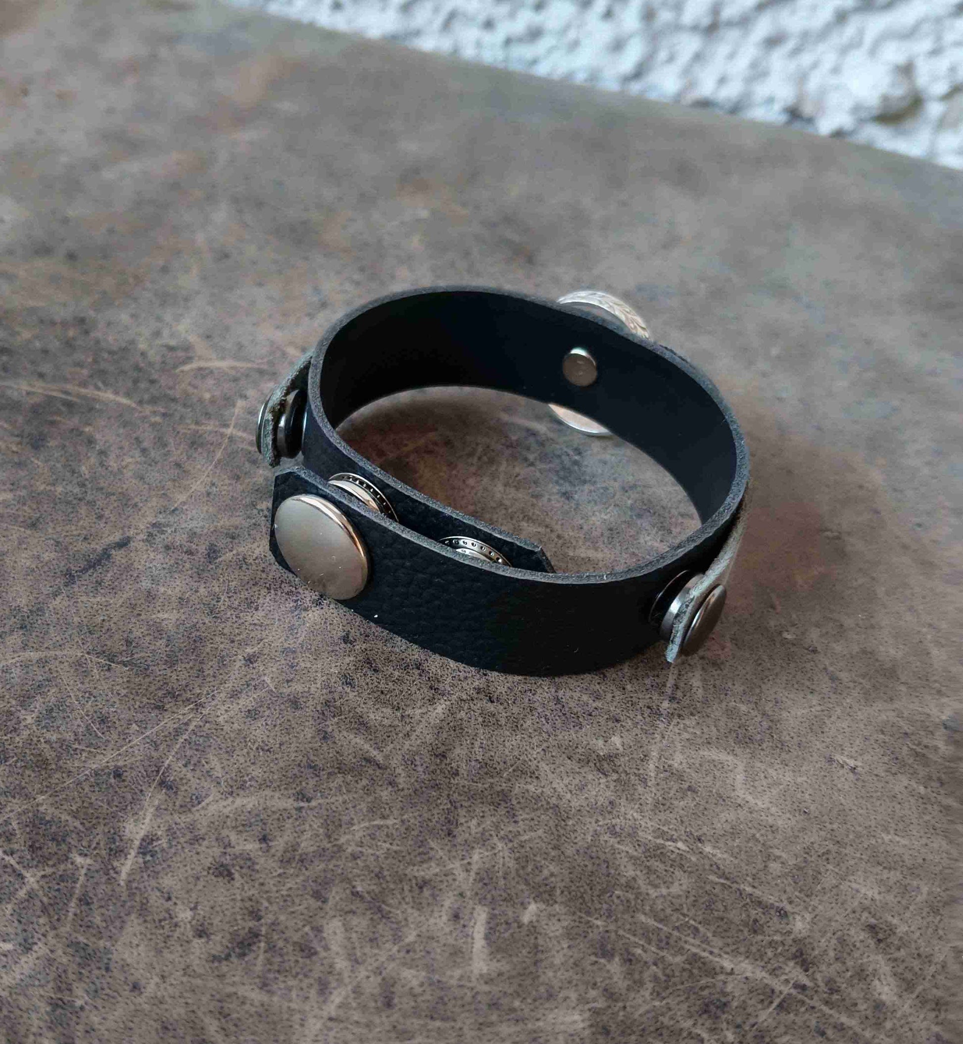 A black textured leather cuff bracelet with silver-toned metal rivets and snap buttons, resting on a textured brown surface.