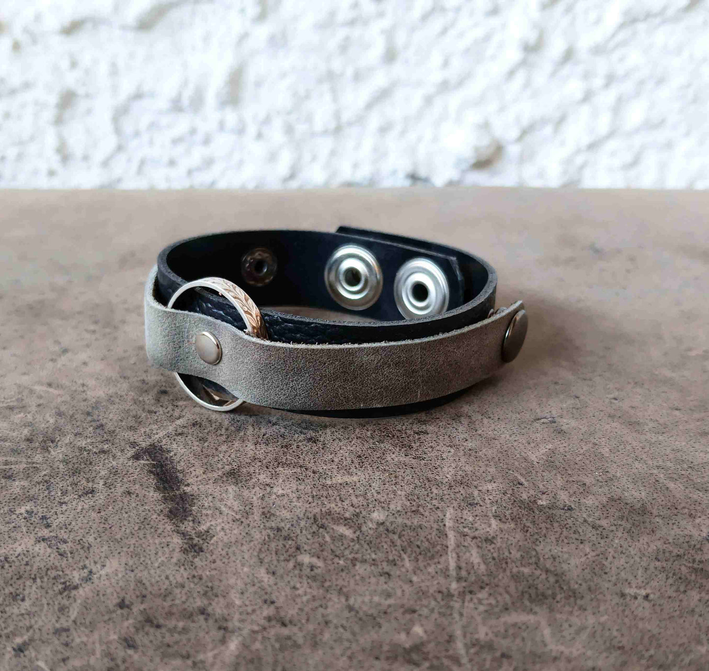 A leather bracelet with a distressed gray-brown outer strap, a black inner lining, and silver-tone metal snap buttons and rings, resting on a textured brown surface.