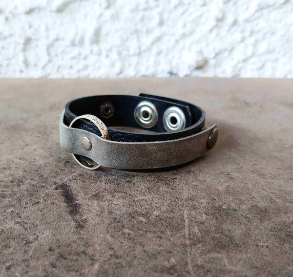 A leather bracelet with a distressed gray-brown outer strap, a black inner lining, and silver-tone metal snap buttons and rings, resting on a textured brown surface.
