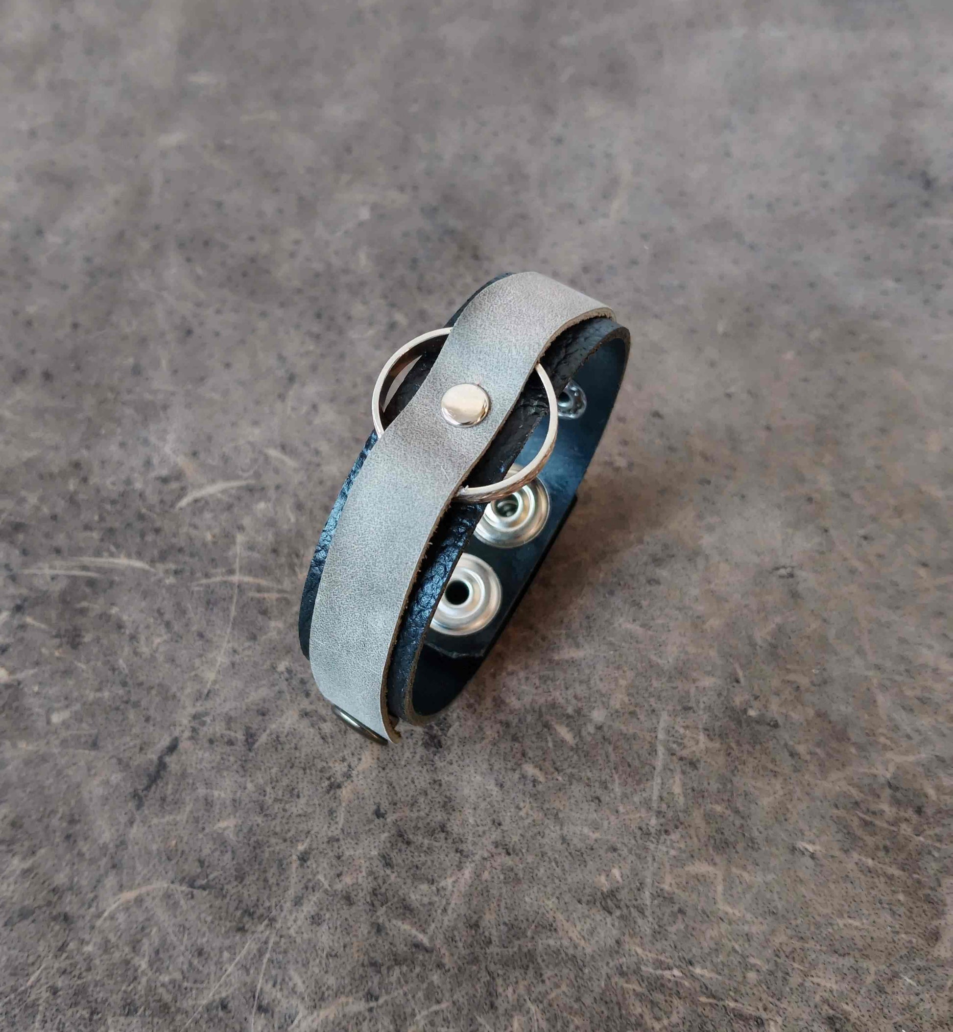A handmade leather wrap bracelet with a light gray suede-like outer band, a dark blue leather inner band, and silver metal grommets and rings, resting on a textured gray surface.