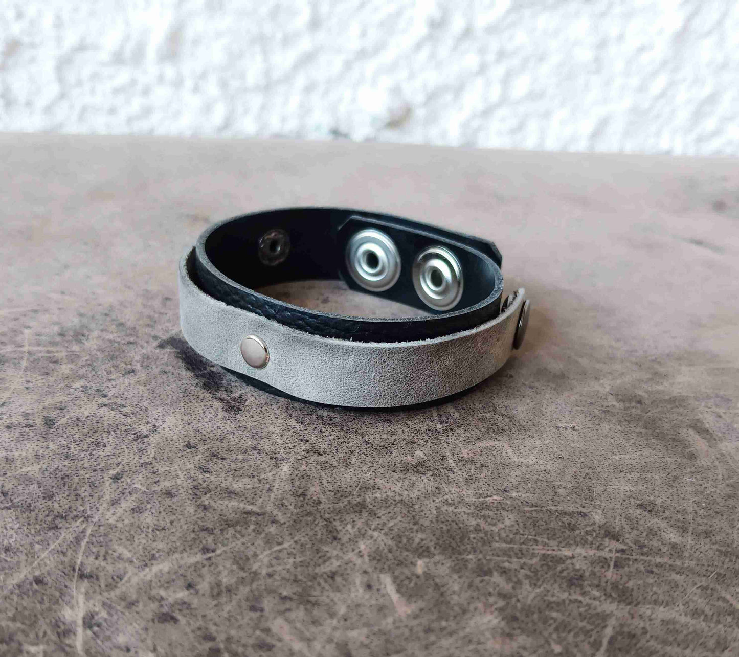 A gray suede leather cuff bracelet with black leather lining and silver metal snap buttons, resting on a textured brown surface.
