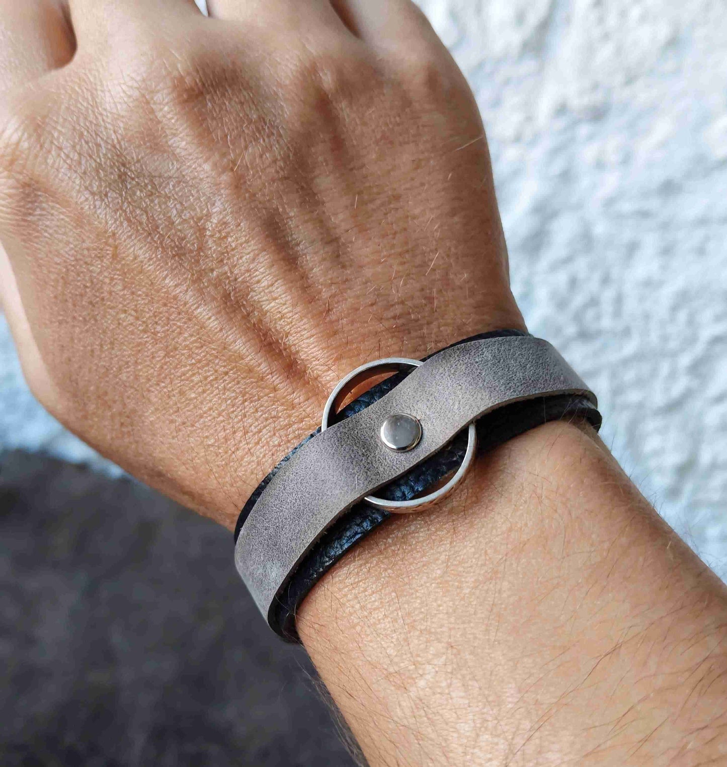 A close-up of a person's wrist wearing a dark gray leather wrap bracelet with a silver-tone metal ring and snap closure.