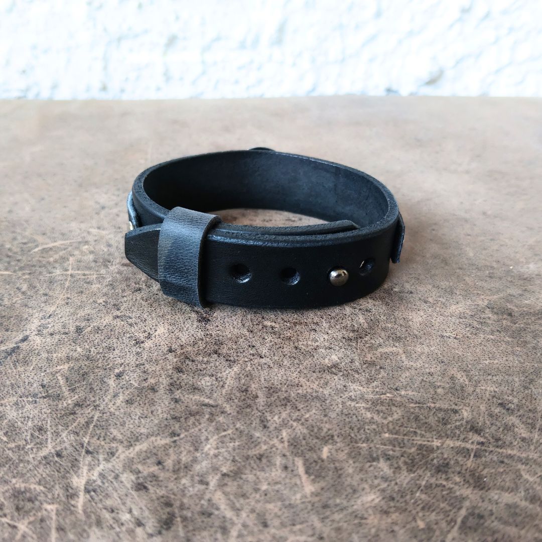A black leather adjustable bracelet with a buckle strap and metal rivets, resting on a textured brown surface.