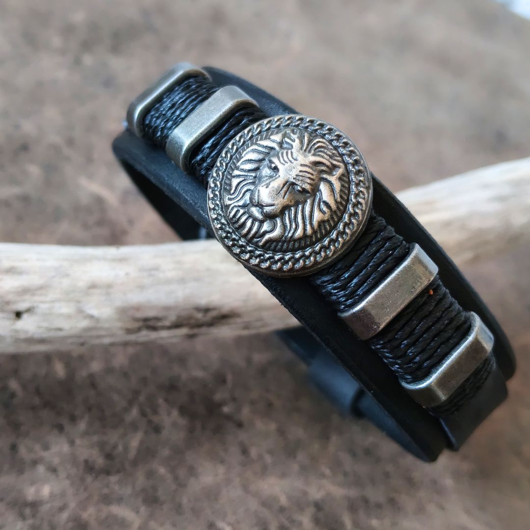 A black leather wrap bracelet with a round antique silver lion head medallion and matching metal accents, displayed on a piece of driftwood over a textured brown surface.