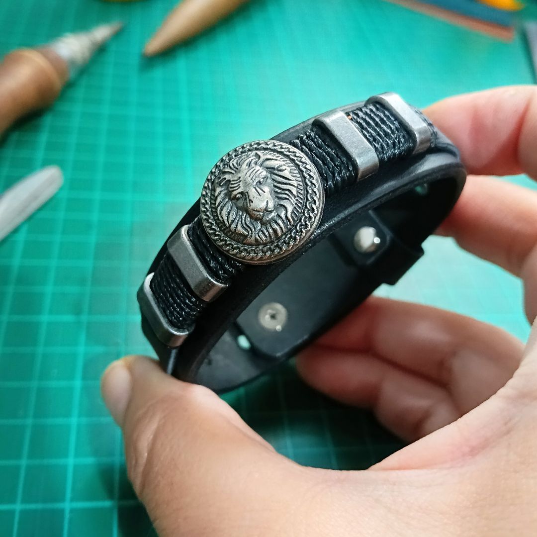 A close-up of a hand holding a black leather bracelet with silver-toned metal accents, including a prominent lion head medallion, set against a green cutting mat background.