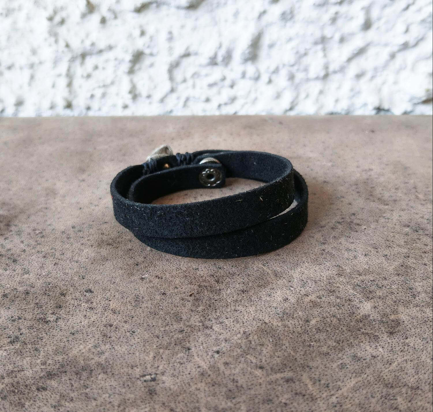 A black multi-strand leather wrap bracelet with a silver-toned metal snap closure, resting on a textured brown surface against a white stucco wall.