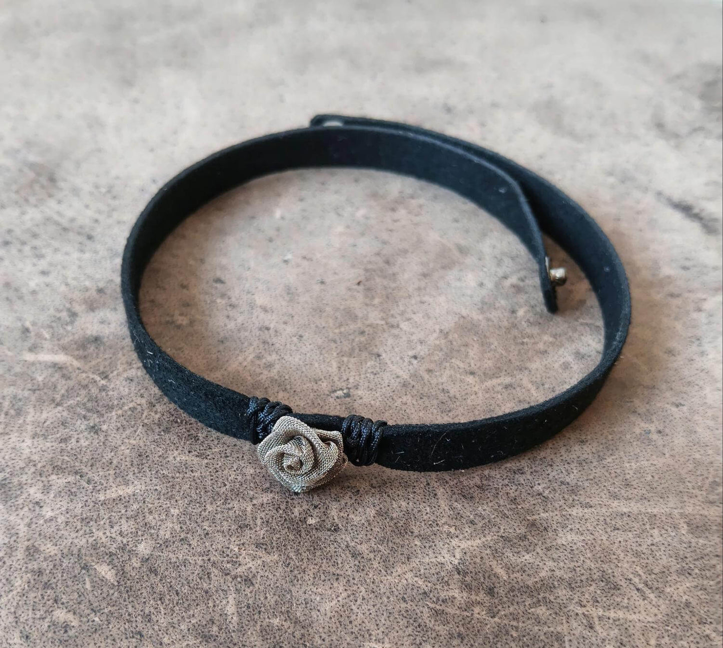 A black suede leather choker with a small silver-toned rose charm, displayed on a textured gray stone surface.