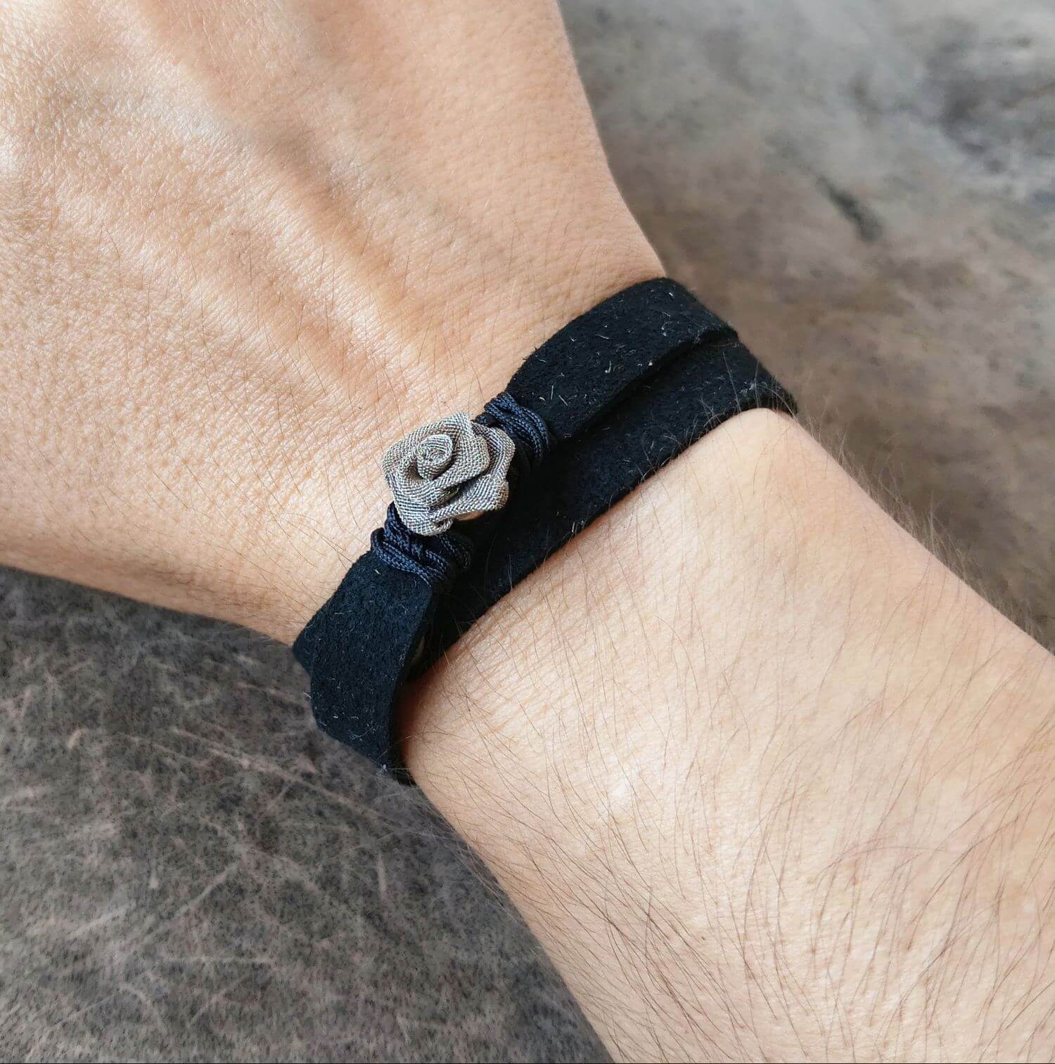 A close-up of a person's wrist wearing a black suede wrap bracelet with a small gray wire rose charm in the center, set against a textured gray background.