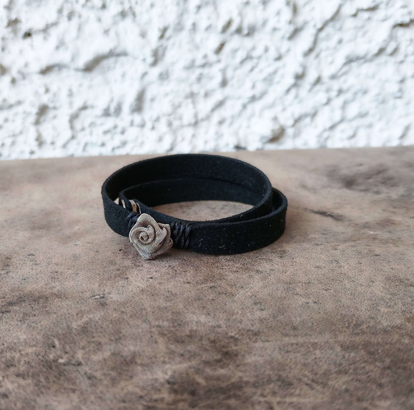 A black wrap bracelet with a small silver-toned rose charm, resting on a textured brown surface against a white stucco wall.