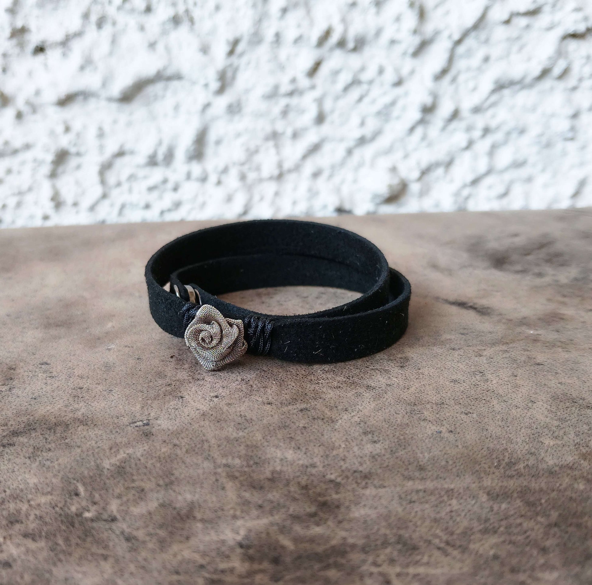 A black wrap bracelet with a small silver-toned rose charm, resting on a textured brown surface against a white stucco wall.