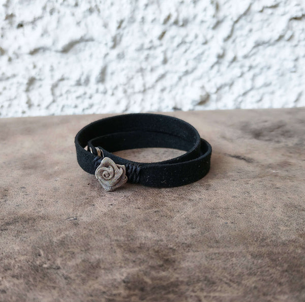 A black wrap bracelet with a small silver-toned rose charm, resting on a textured brown surface against a white stucco wall.