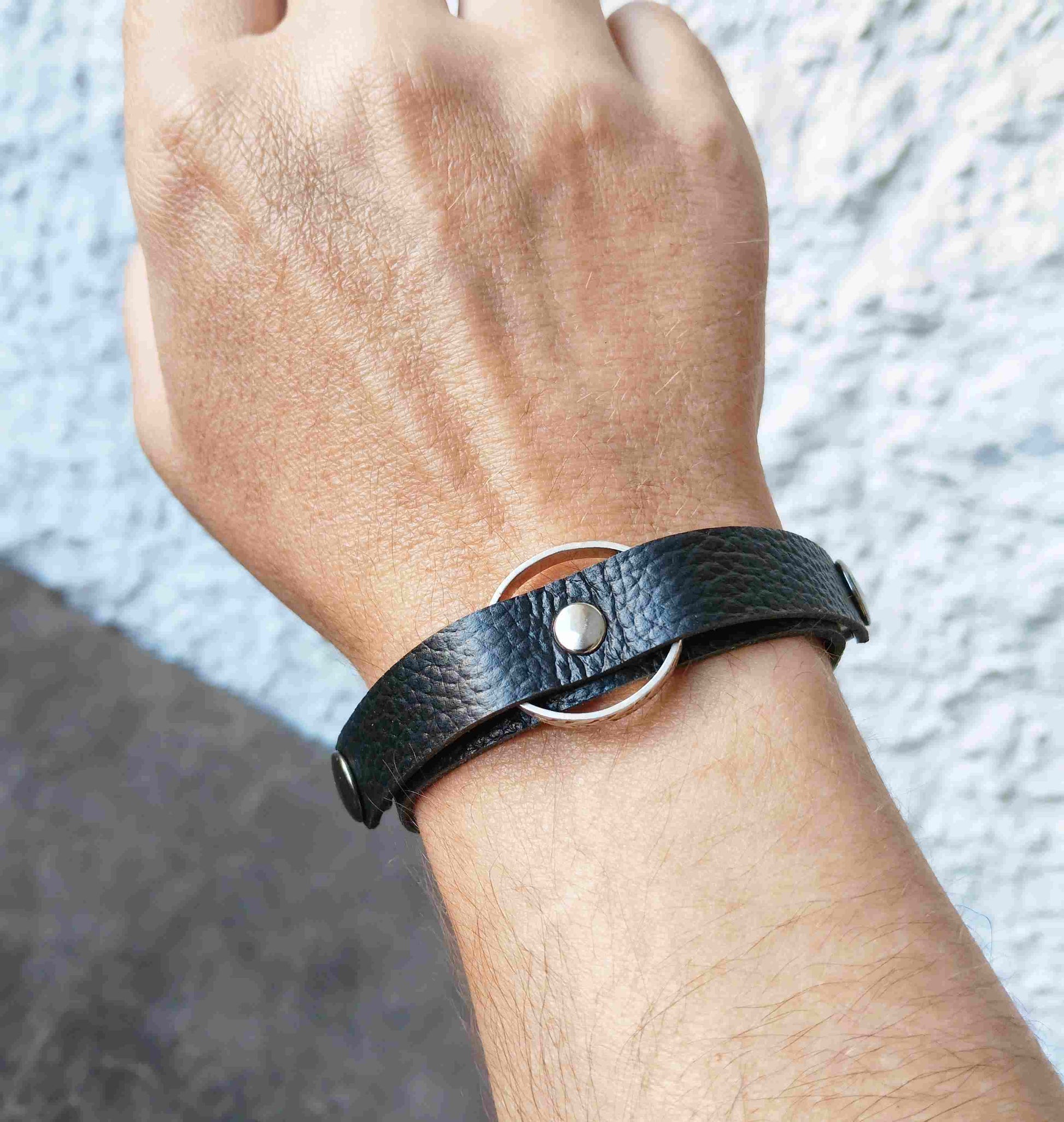 A close-up of a person's wrist wearing a black textured leather wrap bracelet with a silver metal O-ring and snap closure.