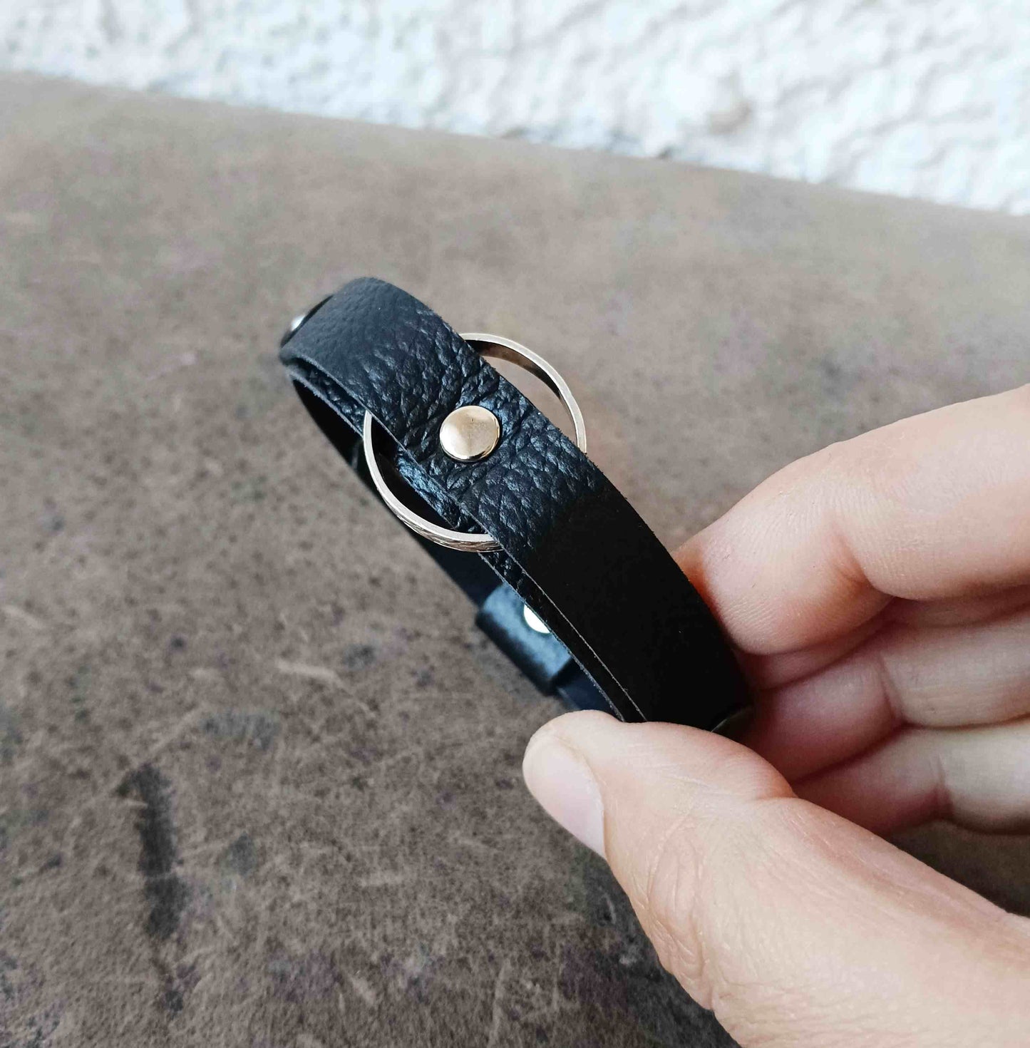 A close-up of a hand holding a black textured leather ring holder bracelet with a silver-tone metal ring and snap button, set against a textured gray surface.