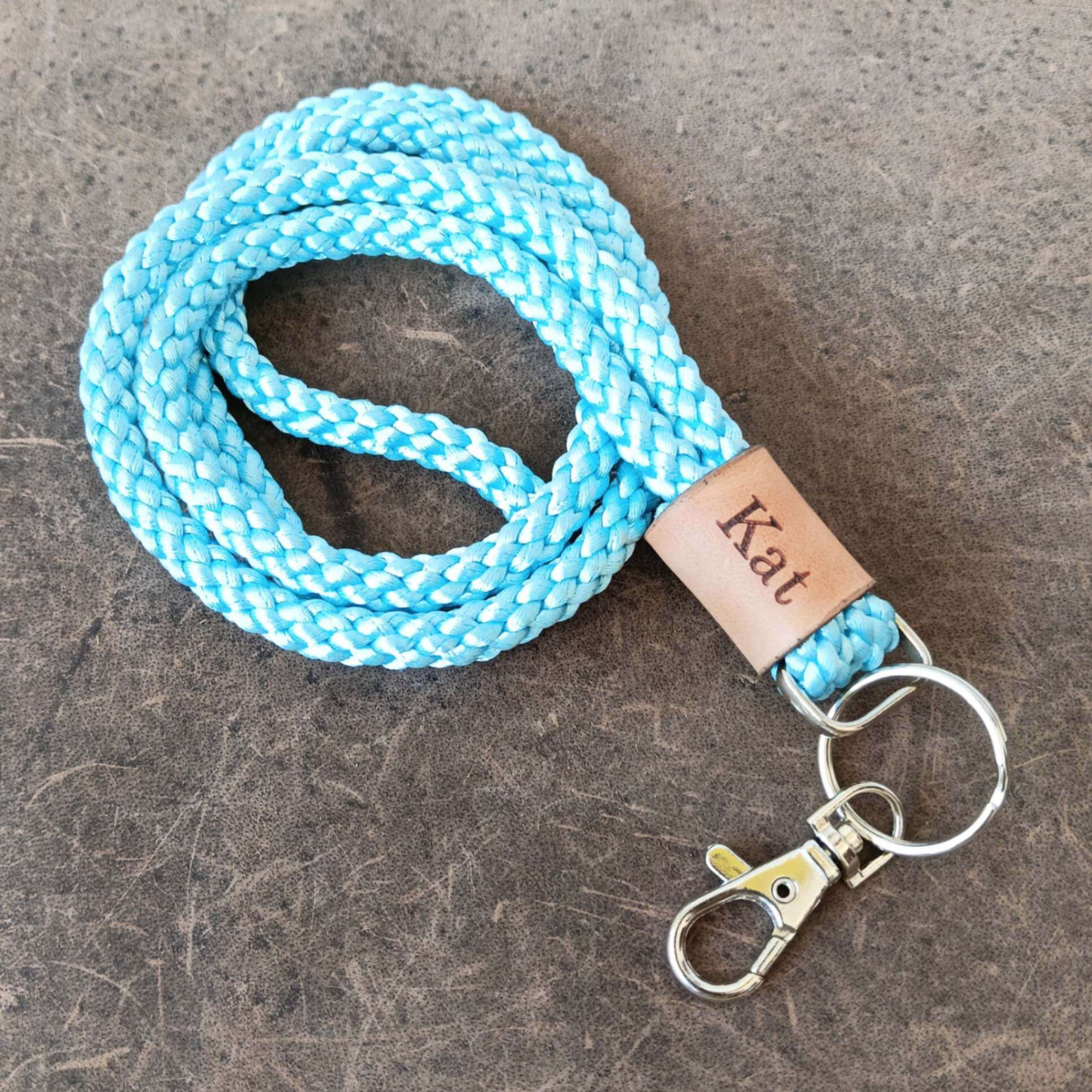 A light blue and white braided rope lanyard with a tan leather tag engraved with the name 'Kat' and silver metal key rings and clasp, displayed on a textured dark gray surface.