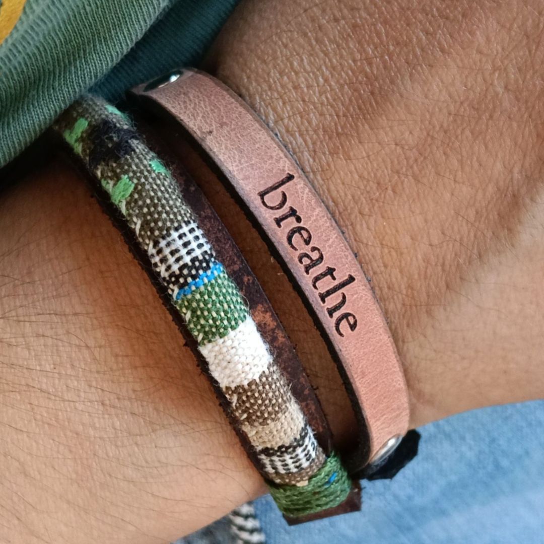 A close-up of a person's wrist wearing a double wrap bracelet: a slim tan leather strip engraved with the word 'breathe' and a woven multicolor fabric bracelet with green, black, white, and brown plaid-like patterns.