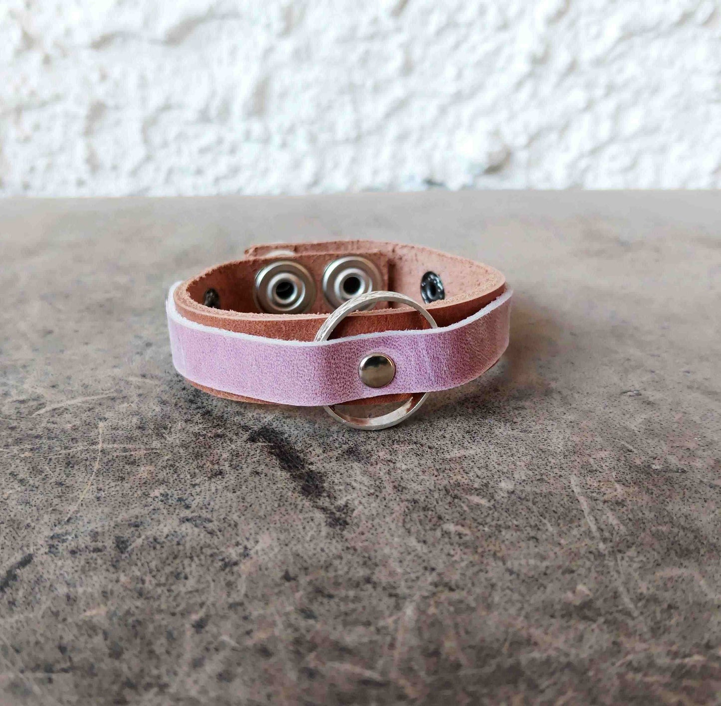 A light pink leather bracelet with a silver metal O-ring and snap button, resting on a textured gray stone surface against a white stucco wall.